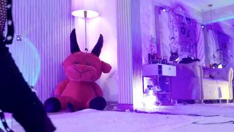 Video of a plush red devil toy with black horns, sitting on a purple carpet, in a dimly lit room with purple and white decor.