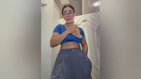 Video of a young woman with fair skin and glasses, wearing a blue crop top and grey pants, standing in a bathroom with beige tiles and a white towel.