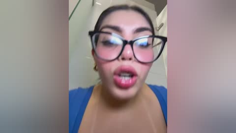 Video of a young woman with fair skin and dark hair, wearing glasses, blue top, and large hoop earrings, with a surprised expression, indoors.