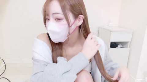 Video of a young Asian woman with fair skin and long brown hair, wearing a white face mask, grey hoodie, and light makeup, adjusting her hair in a minimalistic, softly lit room with white walls and a white dresser.