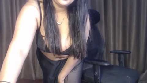Media: Video of a young woman with long black hair, wearing a black lace bra and sheer black pants, leaning over a black office chair, in a dimly lit room with brown curtains.