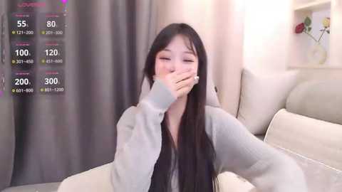 Video of an Asian woman with long black hair, wearing a gray hoodie, covering her mouth, sitting on a beige couch in a cozy room. Weather app with temperature and humidity readings on the left.