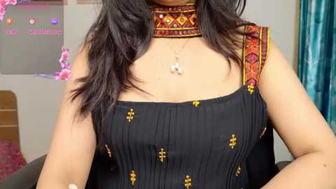 Video of a South Asian woman wearing a sleeveless black dress with gold embroidery and intricate red and gold choker. Background includes a pink floral arrangement, teal curtains, and a white wall with a pink sign.