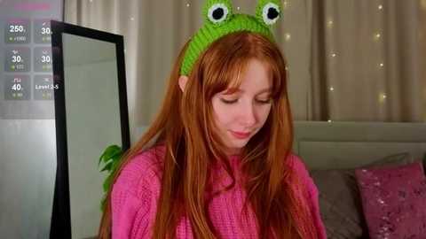 Video of a young, fair-skinned woman with long red hair, wearing a green frog hat, pink sweater, and smiling. Background features a bed with pink pillows and a wall mirror.