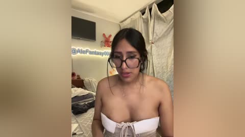 A video of a young woman with long black hair, wearing glasses and a strapless top, standing in a cluttered bedroom.