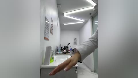 Video of a white-tiled medical examination room with fluorescent lighting, featuring a white sink, a white countertop with bottles, and a hand reaching out from the right.