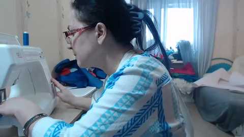 Video of an Asian woman with glasses, dark hair, and red-framed glasses sewing on a sewing machine in a cluttered room with curtains, colorful fabric, and a bed.