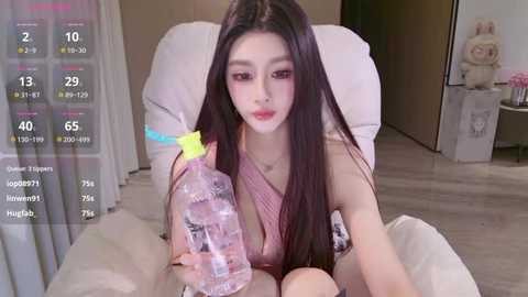 A video of an Asian woman with long black hair, fair skin, and a petite figure, wearing a pink sleeveless dress, sitting on a white couch, holding a clear water bottle with a yellow cap.