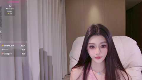 A video of an East Asian woman with long, straight black hair, fair skin, and a slender physique, wearing a pink sleeveless top, sitting on a white pillow in a modern bedroom with beige walls and curtains.