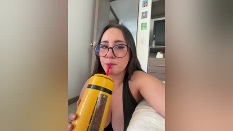 A video of a young woman with glasses, long dark hair, and a black tank top, holding a yellow drink with a red straw, smiling, in a modern bedroom.