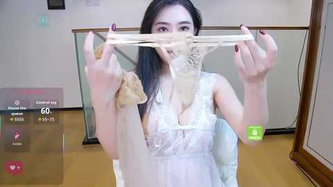 Video of an Asian woman with long black hair, wearing a white lace dress, holding a beige lace bra, indoors, with a virtual overlay showing game stats.