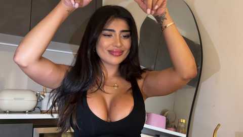 Media: Video of a curvy woman with long black hair, wearing a low-cut black top, raising her arms in a kitchen.