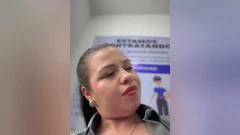 Media: Video of a Latina woman with medium skin tone, brown hair pulled back, wearing a green jacket, looking tired and slightly smiling. Background features a blurred poster in Spanish.