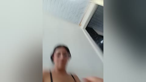 A blurred video of a woman with short dark hair in a black tank top, standing in a small, dimly lit room with a white ceiling and gray walls.