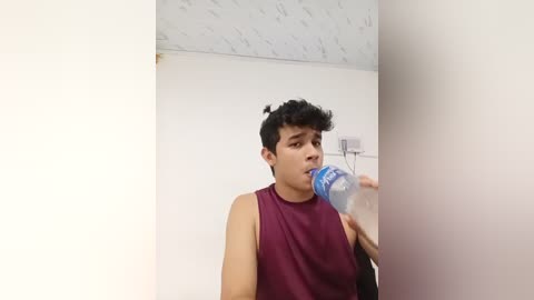 Video of a young man with curly black hair, wearing a sleeveless burgundy shirt, drinking water from a blue bottle, in a white room with a textured ceiling.