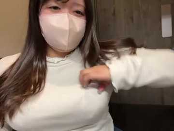 Video of an East Asian woman with long dark hair, wearing a white face mask and a white long-sleeve shirt. She is brushing her hair with her right hand, indoors against a beige wall.