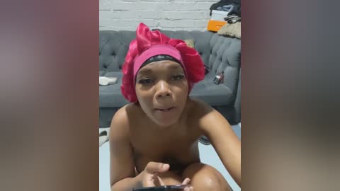 Video of a young Black woman with a large pink turban, topless, sitting on a light blue couch in a modern living room with a gray tufted sofa, white brick wall, and scattered belongings.