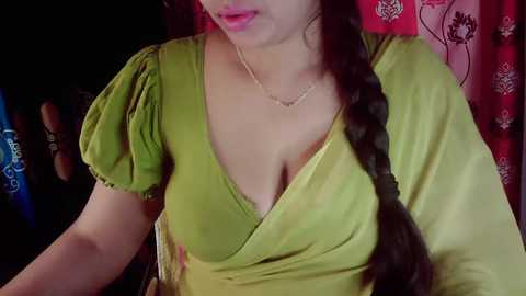 Video of a woman with fair skin, wearing a green blouse with a deep neckline, revealing ample cleavage, and a long, dark braid. Red and blue patterned curtains in the background.