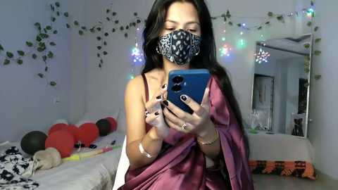 Video of a young woman with long black hair, wearing a black mask and maroon sari, taking a mirror selfie in a cozy bedroom adorned with fairy lights and red balloons.