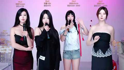 Video of four East Asian women singing into microphones at a promotional event. They wear diverse, elegant outfits, including black strapless dresses, a white blouse, and denim shorts.