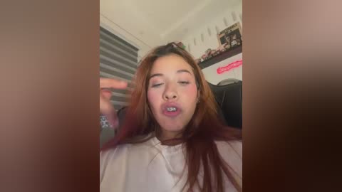 Video of a young woman with long, reddish-brown hair, wearing a white shirt, making a silly face with her tongue sticking out. Background includes a window with blinds, a shelf with decorations, and a gaming chair.
