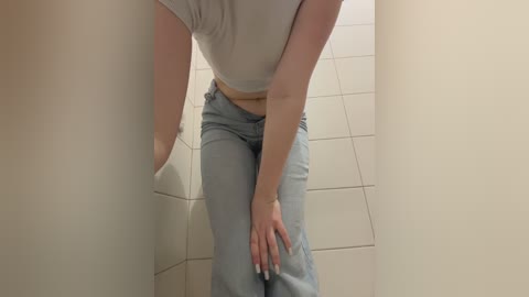 Video of a light-skinned woman with a medium build, wearing a tight white crop top and light blue jeans, sitting on a tiled floor, hands on her knees, in a cramped, beige-walled bathroom.