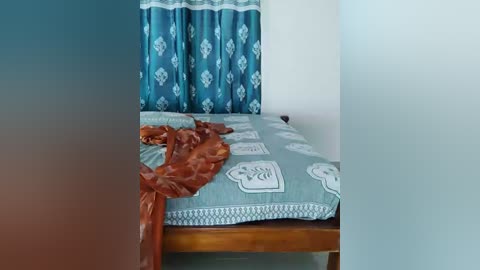 A video of a neatly made bed with a light blue duvet featuring white geometric patterns and an intricately carved wooden headboard. The background features a teal curtain with white diamond patterns. The image is slightly blurred.