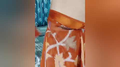 A video of a woman's waist and hips, wearing a vibrant orange skirt with white geometric patterns, standing in a room with teal curtains and a blue-patterned rug.