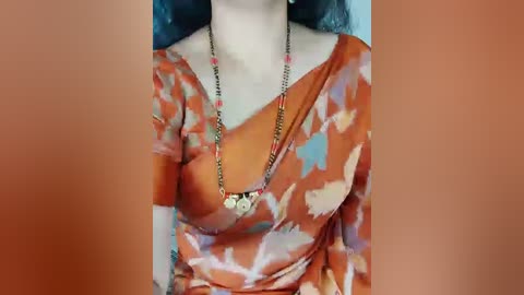 Video of a woman wearing an orange blouse with a floral pattern, adorned with a gold chain necklace featuring small pendants. The background is blurred, focusing on her attire and jewelry.