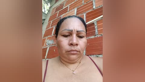 Video of an older, light-skinned Black woman with a round face, wearing a red tank top and gold necklace, standing against a red brick wall with visible mortar.