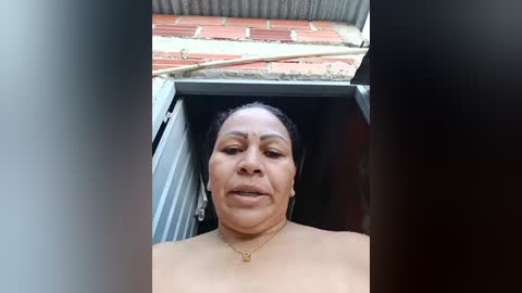 Video of a middle-aged Black woman with a dark complexion, short hair, and wearing a gold necklace, standing inside an open doorway with a red-tiled roof and metal awning visible in the background.