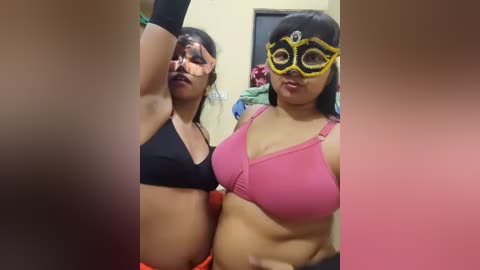 Video of two women, one with black hair and black sports bra, the other with brown hair and pink sports bra, wearing yellow masks, in a room with cluttered background.