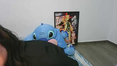 Video of a blue Stitch plush toy resting on a striped blanket, with a framed poster of Iron Man behind it, and a person\u2019s legs partially visible in the foreground.