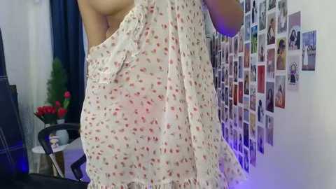 A video of a woman in a floral dress, viewed from the side, standing in a room with a collage of photos on the wall and a table with a vase of red roses.