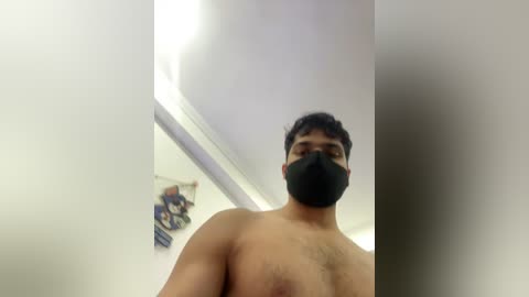 Video of a shirtless man with medium skin tone, dark hair, and a black face mask, taken from a low angle, showing a white ceiling with a small poster and fluorescent lights.