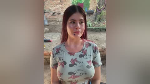 Video of a young woman with straight, dark hair, wearing a Minnie Mouse t-shirt, standing outdoors in a rural setting with a brick wall and greenery.