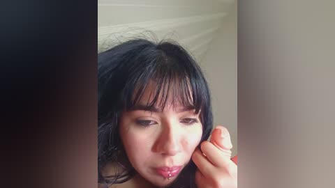 A close-up video of an Asian woman with straight black hair and bangs, wearing light makeup, and a small nose ring, looking down at a hand holding a small object.