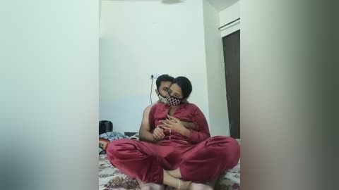 A video captures an Asian couple in matching red pajamas sitting cross-legged on a patterned bed, sharing a tender moment. The man has short black hair, and the woman wears a black mask.