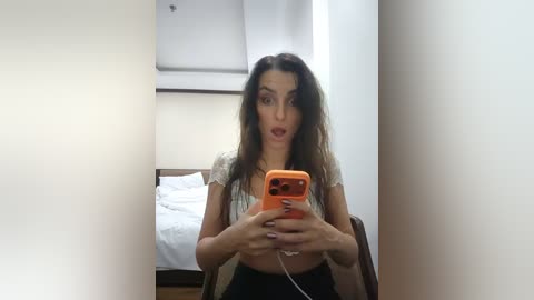 Video of a woman with long, dark hair, wearing a white top, and black pants, taking a selfie in a bedroom with a white bed and beige walls.