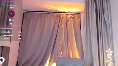 Video of a modern bedroom with soft, warm lighting. A large window with sheer, pink curtains is partially open, revealing a cozy, sunlit interior. A white ceiling fan is visible in the background.