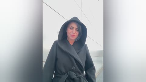 Video of a woman in a black trench coat with hood, standing against a white wall, wearing a subtle smile.