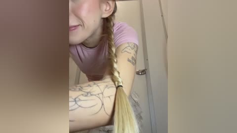 Media: Video of a smiling woman with braided blonde hair, wearing a light purple shirt, leaning over a railing. She has tattoos on her left arm.