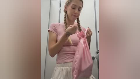 Media: Video of a young, fair-skinned woman with blonde braided hair, wearing a pink short-sleeved top and white skirt, holding a pink plastic bag in a public restroom with white stall doors.