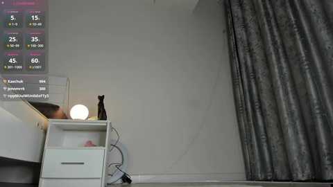 Video of a minimalist bedroom with a gray curtain, white nightstand, and a black cat statue; digital weather app display in the top left corner.