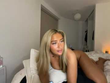 Video of a blonde woman with long hair, wearing a white tank top, lying on a bed in a modern, minimalistic bedroom.
