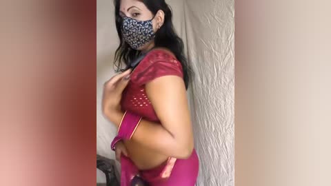 A video of a dark-skinned woman with long black hair, wearing a red saree, face mask, and gold bangles, posing indoors.