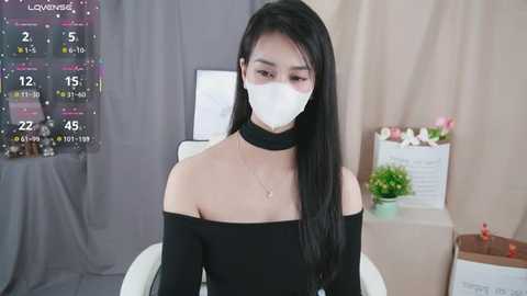Video of an Asian woman with long black hair and fair skin, wearing a black off-shoulder top, white mask, and choker necklace. Background features a calendar, flowers, and a gray curtain.