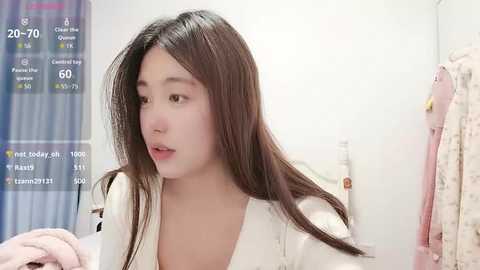 Video of a young East Asian woman with long black hair, wearing a white top, standing in a minimalist bedroom with a pink blanket and white curtains.