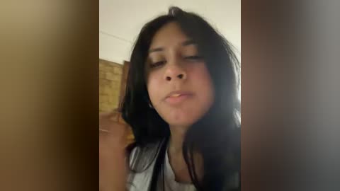 A close-up video of a young woman with long black hair, medium complexion, and closed eyes, making a kissing gesture. She wears a black jacket over a white shirt, against a beige wall with a wooden frame in the background.
