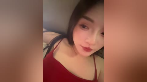 A video of an East Asian woman with fair skin, long black hair, and red lipstick, wearing a red spaghetti-strap top. She has a neutral expression. The background is blurry, focusing on her face.